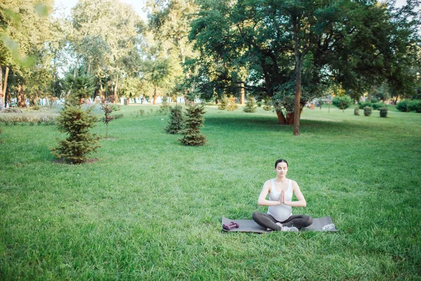Calm and peaceful pregnant woman sit in lotus pose in park outside alone. She hold hands together in pray position. Model looks calm and peaceful.