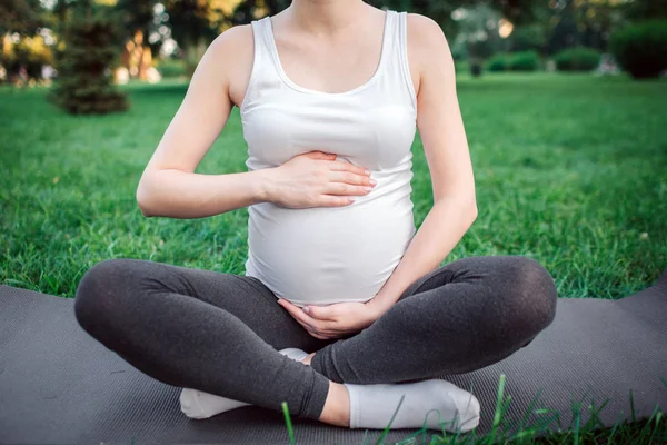 Nacięcie młoda kobieta w ciąży siedzieć na Mat Joga w parku. Ona trzymać się za ręce wokół brzucha. Model siedzieć w pozycji lotosu. — Zdjęcie stockowe