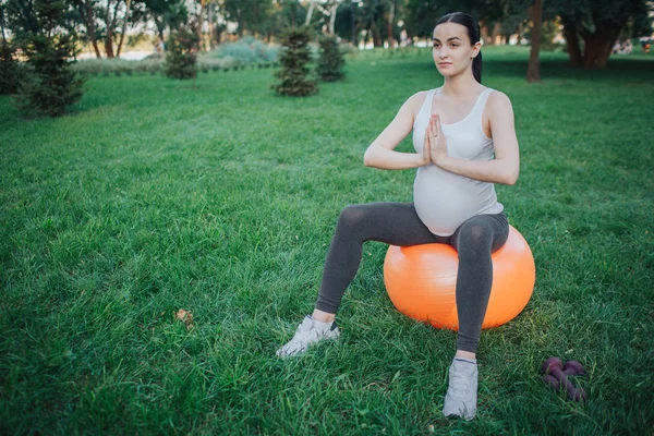 Mujer embarazada joven sentarse en la pelota naranja fitness en el parque verde y meditar. Ella toma de la mano en pose de oración . —  Fotos de Stock