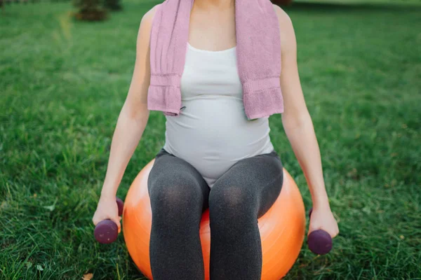 Wyciąć widok młodej kobiety w ciąży, siedzi na piłka pomarańczowy fitness w parku. Ona trzymaj hantle w ręce i ręcznik wokół szyi. — Zdjęcie stockowe