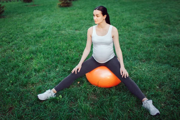 Jovem mulher grávida sentar-se na bola fintess laranja com pernas espalhadas no parque. Ela olha para a direita . — Fotografia de Stock