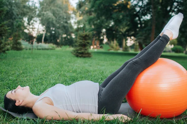 Bonita joven embarazada acostada en yoga mate en el parque. Ella sostiene las piernas en la pelota naranja fitness y mira hacia arriba. Modelo estirar hacia atrás y todo el cuerpo . — Foto de Stock