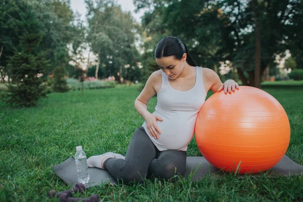 Linda joven embarazada sentada en yoga mate en el parque. Ella toma de la mano en el vientre y lo mira. Modelo inclinado a bola naranja fitness . —  Fotos de Stock