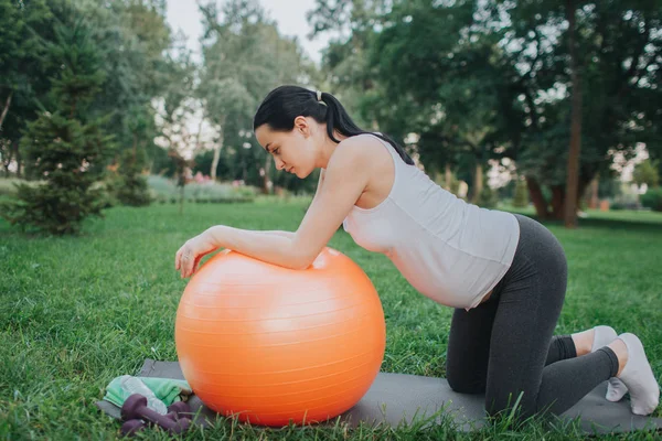 Linda joven embarazada haciendo ejercicio en el parque. Se pone de rodillas y mira hacia abajo. Modelo magra a gran bola naranja fitness . — Foto de Stock