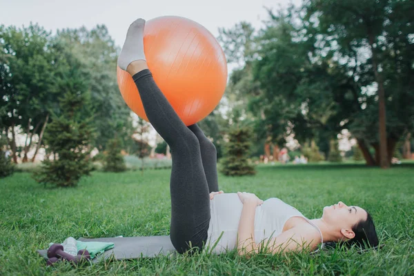 Mujer embarazada joven concentrada ejerciendo sobre yoga mate en el parque. Ella sostiene una gran pelota naranja de fitness entre las piernas. Modelo mantener las manos en el vientre . — Foto de Stock