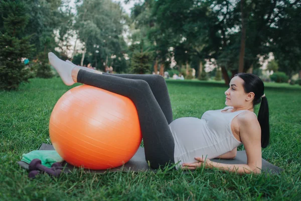 Jovem grávida sofre de dor na área dos quadris. Ela está deitada no ioga mate no parque. Modelo mantenha as mãos nos quadris e pernas na bola laranja . — Fotografia de Stock