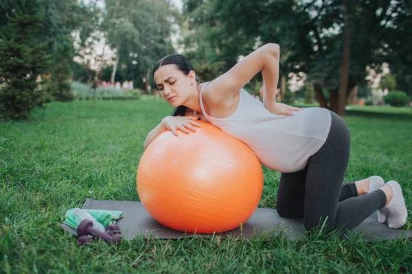 Mladá těhotná žena suffeer od bolesti zad. Ona stojí v kolenou v parku. Model štíhlá, oranžové fitness míče. — Stock fotografie