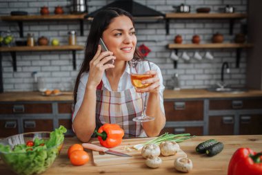 Genç kadın oturup mutfakta masada. Beyaz şarap nd konuşma telefonda içki. Kadın mutlu görünüyor. Masanın üzerinde yatan sebze.