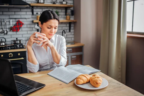 Intelligent adult woman sit at table in kitchen. She read journal and hold white cup with hot drink. Woman look down. She work at home.