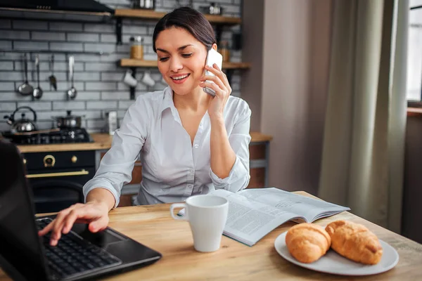 Nette, fröhliche Frau sitzt am Tisch und arbeitet zu Hause. Sie tippt auf der Laptop-Tastatur und telefoniert. Ihr Frühstückstisch steht auf dem Tisch. — Stockfoto