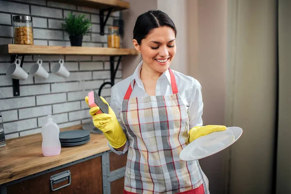 Mulher agradável no suporte de avental na cozinha e posar. Ela segura a placa branca e sorri. Limpador feminino profissional em luvas amarelas . — Fotografia de Stock