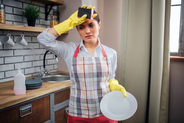 Limpador fêmea cansado ficar na cozinha e segurar a mão na testa. Ela usa avental e luvas amarelas. Mulher segurar placa branca . — Fotografia de Stock