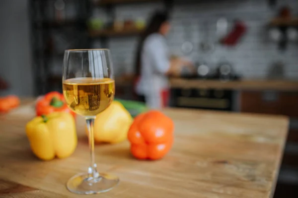 Glas vitt vin står på skrivbord med gula och orange paprika. Kvinna står vid spisen i köket och koka. — Stockfoto