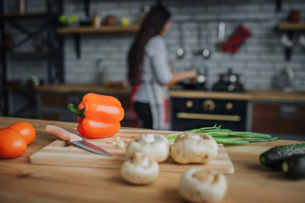स्वयंपाकघरात डेस्क टेबलवर भाज्या आणि चाकू बंद करा. बाई स्टोव्हवर उभी राहून स्वयंपाक करतात. ती व्यस्त दिसते . — स्टॉक फोटो, इमेज