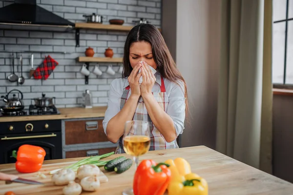 Νεαρή γυναίκα να καθίσει στο τραπέζι στην κουζίνα και snezing. Έπιασε κρύο. Άρρωστος μοντέλο κάλυψη στόμα woth χαρτοπετσέτας. Λαχανικά που βρίσκονται στο τραπέζι. — Φωτογραφία Αρχείου