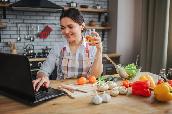 Nice cheerful woman sit at table in kitchen. She type on keyboard laptop and smile. Model hold glass of white wine. Vegetales lying on table.