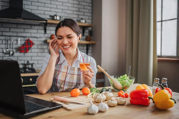 Neşeli, güzel, çekici bir kadın mutfaktaki masada oturuyor. Gülümsedi ve kulaklıklara tel kulaklık taktı. Dizüstü bilgisayarda model görünümü. Sebzeler masada yatıyor..