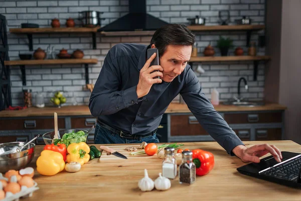 Adult businessman stand at kitchen table desk and reach laptop keyboard. He talk on phone. Guy is concentrated. Colorful vegetables lying on desk.
