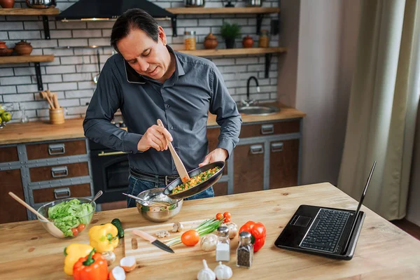 Zaměstnaný člověk stojí u stolu v kuchyni a směs vajec. Mluvit na telefonu a podívejte se. Barevné zeleniny, ležící na stole. — Stock fotografie