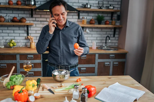 Busy man stand at table in kitchen and talk on phone. He smile and hold egg in hand. Guy cooking. Colorful vegetables on table.