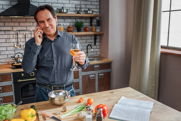 Happy adult man stand at table in kitchen and talk on phone. He hold glass of white wine and smile. Man cooking. Vegetalbes and cookbook on table.