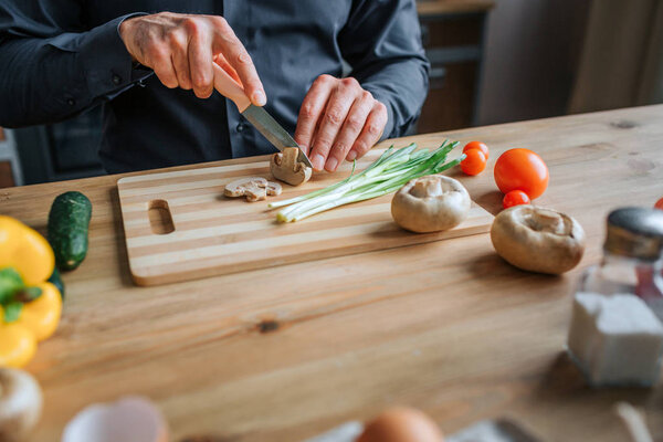 Cut view of man hands cutting mushrooms with knife. He work with both hands. Man stand in kitchen at table.