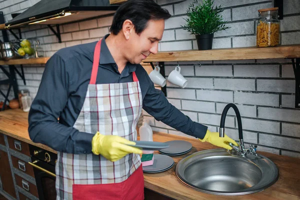 Bom homem em apon stand na cozinha e toque pia. Ele segurou o prato. O tipo usa luvas de lavar . — Fotografia de Stock