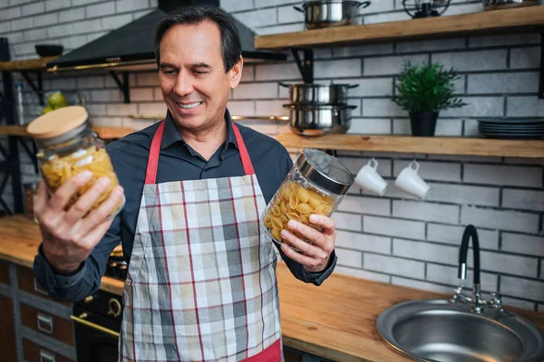 Man in apron stand in kitchen and hold two jars of spagetti. He look at them and smile. Man stand alone.