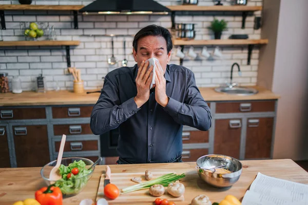 Dospělý nemocný kýchání na bílém ubrousku. On stojí u stolu v kuchyni. Stůl plný barevných zdravé zeleniny a koření. — Stock fotografie