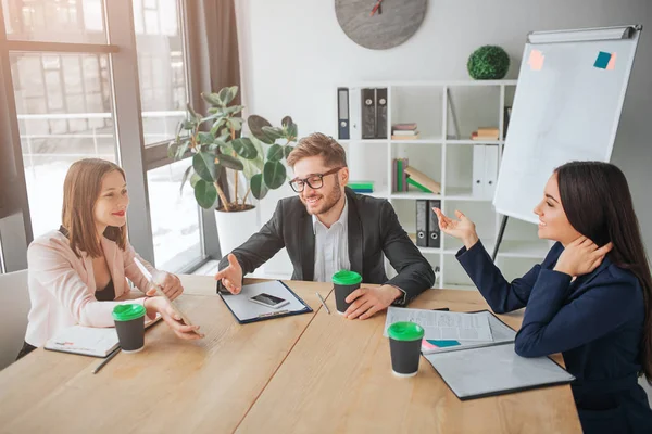 Bonitos hombres y mujeres jóvenes se sientan juntos en la mesa en la sala de reuniones. Tienen conversación y se ríen. La gente trabaja y habla . — Foto de Stock