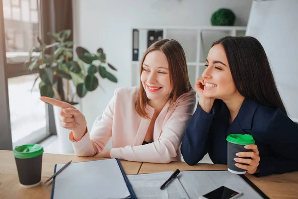Twee jonge vrouwen zitten samen aan tafel in mooeting kamer. De punt van een model met vinger en lachen. Haar vriend houd kopje koffie en kijk naar links. Ze lachen ook. — Stockfoto