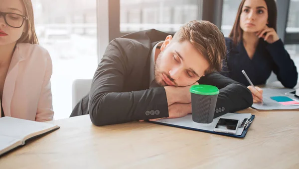 Tired young man sit at table and sleep in meeting room. He hold head on hands. Young women sit beside him and listen. They look concentrated. — Stock Photo, Image