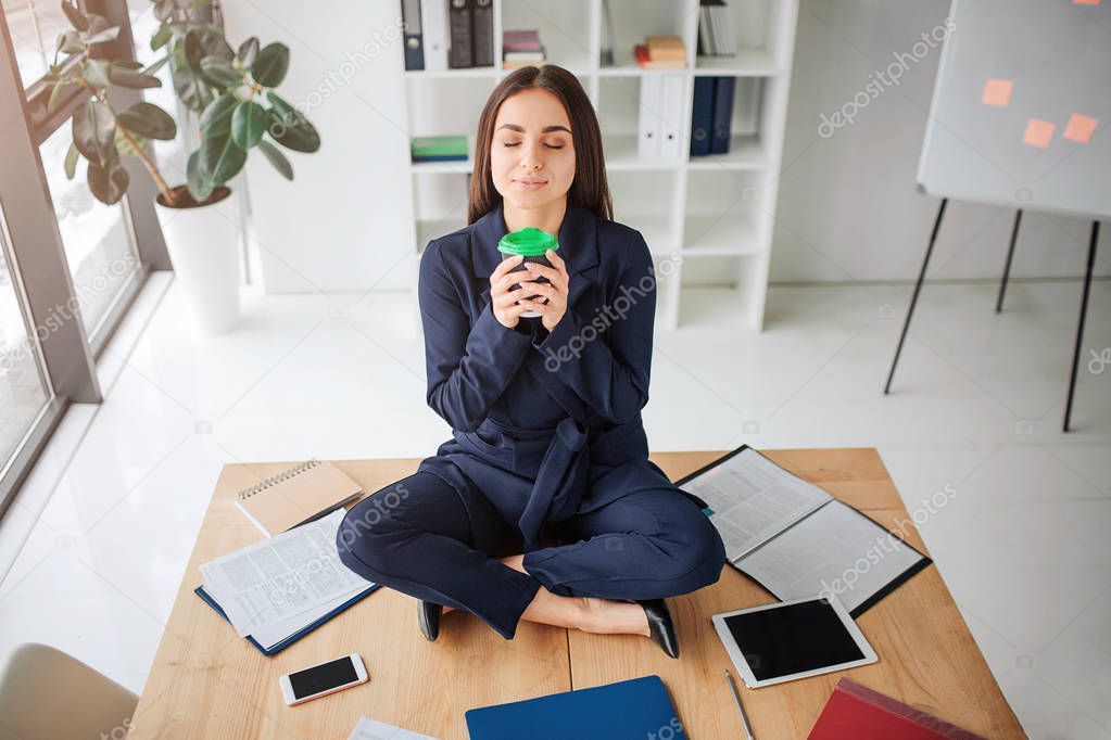 Calm peaceful young woman sit on table in lotus pose in room. She hold cup of coffee and smell it. Young woman enjoy. Materials and electronics on table.