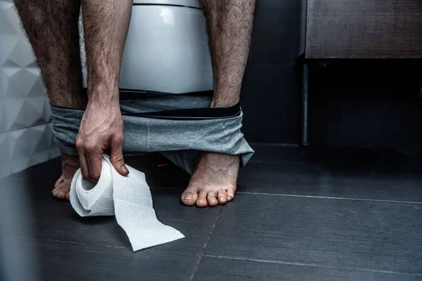 Vista panorâmica. O homem senta-se na sanita e alcança o papel. As pernas são finas e pálidas. Problemas com o estômago. Obstipação. Reação na cama. Sofrer . — Fotografia de Stock