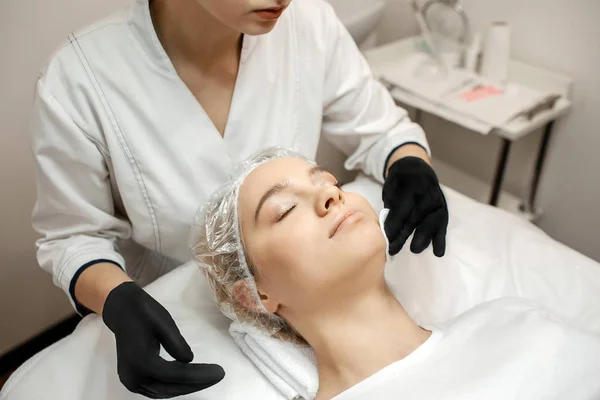 Mujer joven con gorra acostada en el sofá en la habitación. esteticista de pie a su cabeza y haciendo procedimientos con las manos. Disfruta y relájate . — Foto de Stock