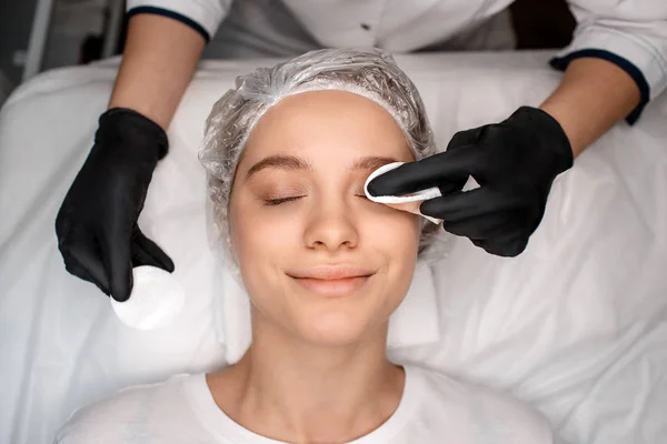 Mujer joven y feliz tumbada en un sofá blanco en la sala de esteticistas. Cosmetólogo utilizar esponja para párpados cuidado de la piel. Cuidado con el trabajo. Disfruta. . — Foto de Stock