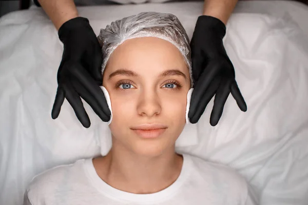 Calm peaceful young woman lying on black couch. Beauticians hands in black gloves holding white sponges and touching clients skin.
