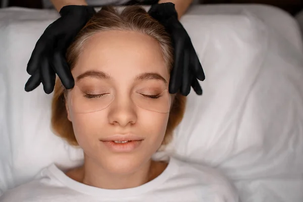 Mujer joven y relajada disfrutando de masajes en la cabeza. Ella mantiene los ojos cerrados. Las manos en guantes de látex negro tocan los lados de su frente . — Foto de Stock