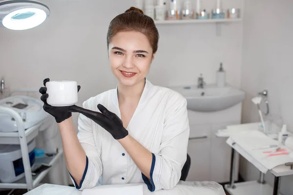 Belle jeune esthéticienne tenir pot de crème pour le visage. Elle regarde la caméra sourire et pose. Modèle assis dans la chambre. Elle pointe sur le pot de crème . — Photo