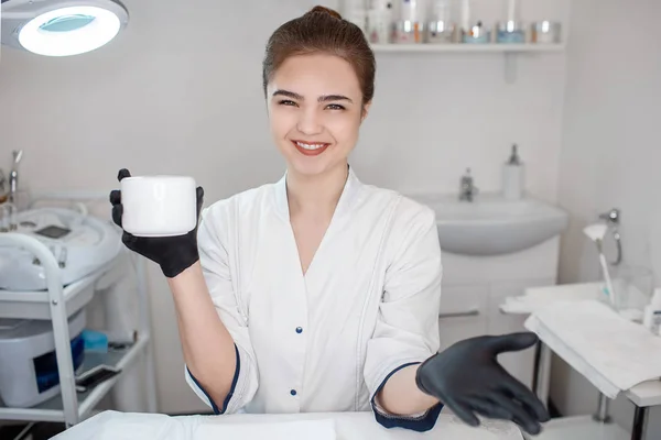 Attrayant jeune esthéticienne tenir pot de crème visage et sourire. Elle pose à la caméra dans la salle de cosmétologie . — Photo