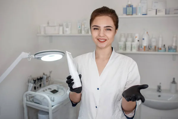 Joyeux beau stand esthéticienne dans la salle de cosmétologie et tenir la crème tube. Elle regarde la caméra et sourit . — Photo