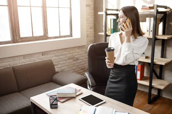 Ung affärskvinna monter på bordet i rummet. Hon prata telefon och titta på fönstret. Modell hålla kopp kaffe. — Stockfoto