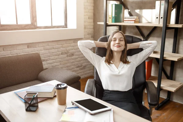 Ung kvinna med resten. Hon sitter vid bordet i rummet med slutna ögon. Modellen leende. hon håller händerna bakom huvudet. — Stockfoto