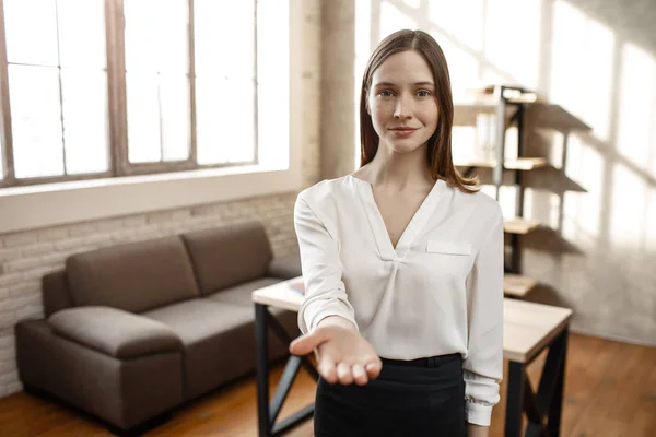 Ung affärskvinna nå hand till kameran. Hon ser raka och ler lite. Tomma rum. Ensam. Dagsljus. — Stockfoto