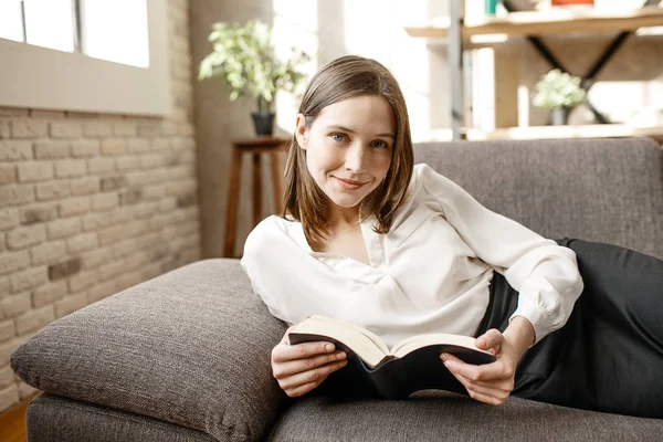 Bella giovane donna d'affari sdraiata sul divano in camera. Lei posa sulla macchina fotografica e sorride. Donna tenere aperto libro . — Foto Stock