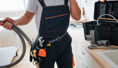 Mutfakta ayakta ve hortum tutan adam görünümünü kesti. O çalışma üniforma giyiyor. Kemer araçlarda. Lavabo standında adam.