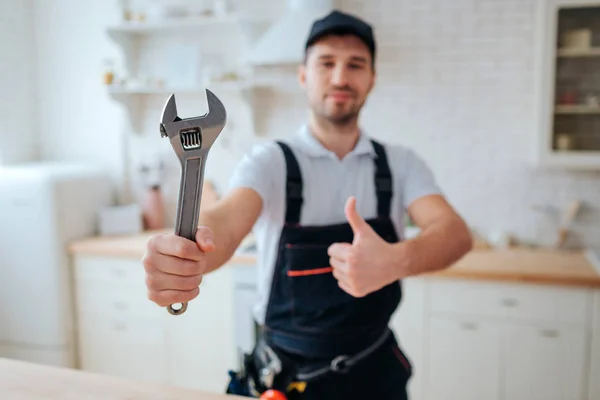 Young plumber hold wrench in hand. He show it to camera and hold big thumb up. Guy stand in kitchen. White background. Daylight.