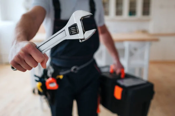 Cut view of handyman holding tool box and wrench in hands close to camera. He stand in kitchen. Guy wear special uniform. Daylight.