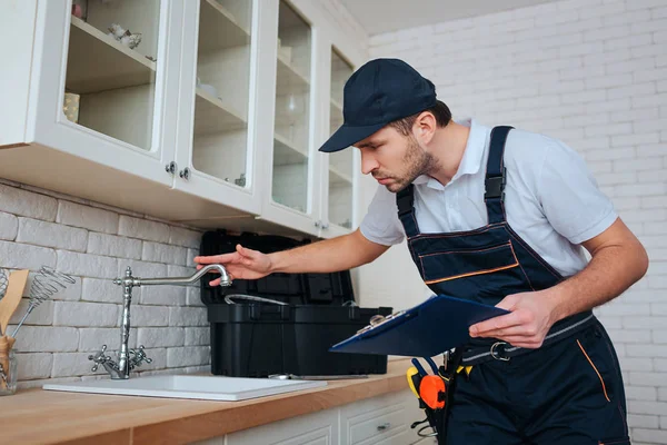 Careful young plumber stand in kitchen and check water tap on sink. He hold plastic tablet in hand. Serious and concentrated. — Stock Photo, Image