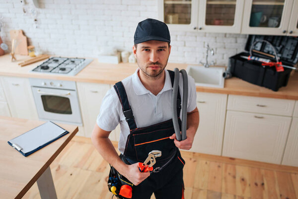 Confident calm young handyman look on camera. He stand in kitchen and hold wrench. Plastic tablet with paper on table. Toolbox on desk.